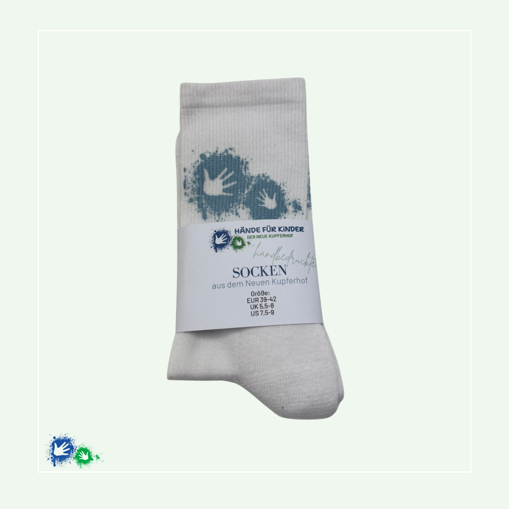 Zusammengefaltete weiße Socken mit hellblauem Hände-Logo. Darum eine Papier-Banderole mit dem Aufdruck: Hände für Kinder Logo, handbedruckte Socken aus dem Neuen Kupferhof, Größe: EUR 39-42, UK 5,5-8, US 7,5-9